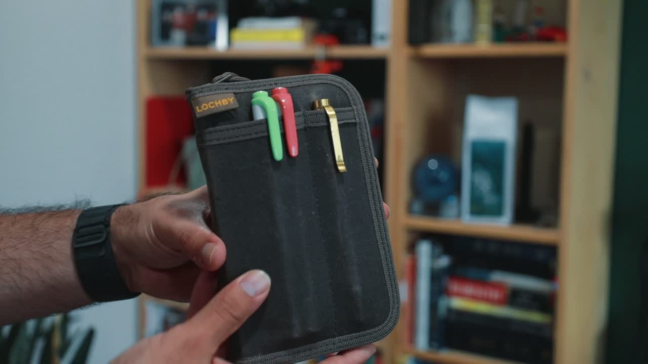 LOCHBY Pocket Journal. Dark waxed canvas LOCHBY Pocket Journal held in hand, brass leather LOCHBY tab, front elastic loops holding two highlighters and a brass clip; opens to the mustard interior and kraft LOCHBY notebook.