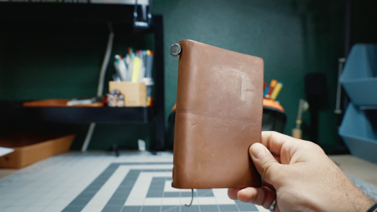 Traveler's Company Notebook (Passport, Camel). Camel leather Traveler's Company Passport Size notebook held in one hand, closed, turned slightly, elastic closure visible on the spine.