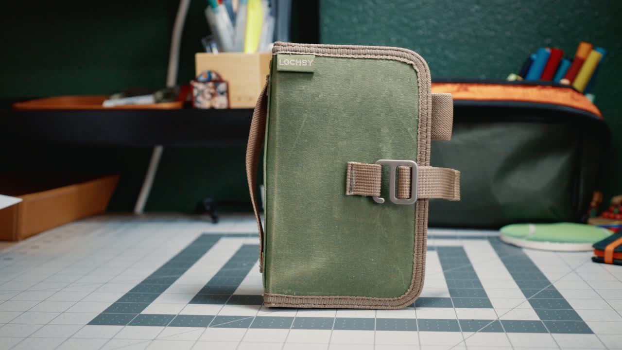 LOCHBY Mini Field Journal. Olive waxed canvas LOCHBY Mini Field Journal standing upright on the cutting mat, tan webbing strap and metal buckle across the front, LOCHBY tab top left.